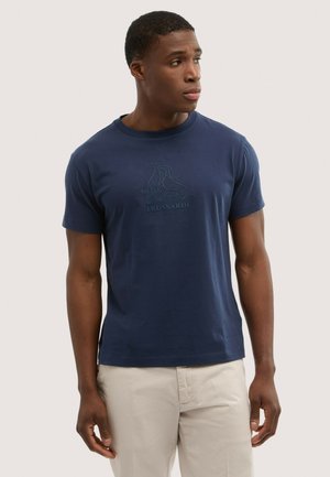 Uomo che indossa una T-shirt Trussardi blu navy con logo in rilievo e pantaloni beige, che guarda verso destra su uno sfondo neutro.