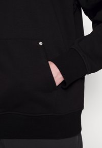 Sudadera negra con un bolsillo frontal, que cuenta con un detalle de botón a presión plateado. Textura suave con puños y dobladillo de canalé. No se ven gráficos ni patrones.