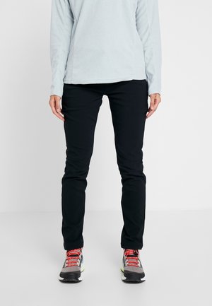 Persoon draagt zwarte slim-fit broek, lichtgrijs shirt met lange mouwen en grijze wandelschoenen met rode veters, staand op een witte vloer.