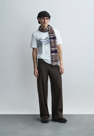 Λευκό κοντομάνικο μπλουζάκι με γραφικό ζιγκ-ζαγκ σε μπλε και γκρι, συνδυασμένο με καφέ παντελόνι φαρδιάς γραμμής και πολύχρωμο πλεκτό κασκόλ.