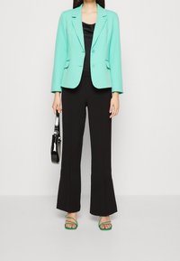 Kvinna iklädd en mintgrön blazer, svart topp, svarta byxor, gröna sandaler och håller en svart handväska framför en plain vit bakgrund.