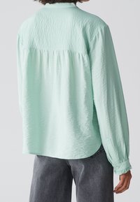 Camicia verde menta con tessuto testurizzato, maniche lunghe e schiena arricciata. Presenta polsini con bottoni e un orlo arrotondato.