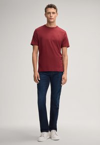 Bordeauxfarbenes Crew-Neck-T-Shirt aus Baumwolle, kombiniert mit dunkelblauen Jeans. Das Modell trägt weiße Sneaker. Einfaches Design, kurze Ärmel, keine Muster.