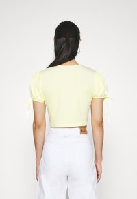 Gul cropped top med puffede ærmer og bindedetaljer. Fremstillet i blødt stof. Modellen bærer hvide bukser med høj talje.