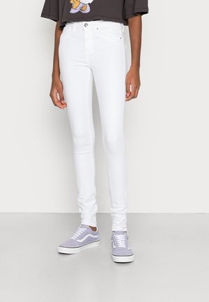 Personne portant un jean blanc slim, des baskets grises et blanches, et un t-shirt court foncé, debout contre un mur uni.