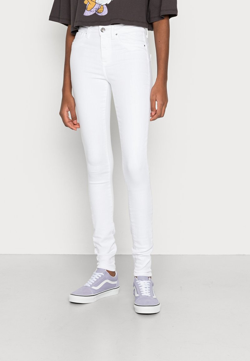 Personne portant un jean blanc slim, des baskets grises et blanches, et un t-shirt court foncé, debout contre un mur uni.