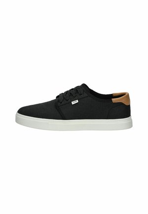Zapatillas de lona negras con suela de goma blanca y acento de cuero marrón en el talón. Cuentan con cierre de cordones y un pequeño logotipo en el costado.