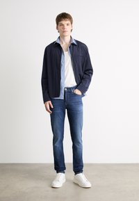 Camisa arrugada azul marino sobre una camisa de botón azul claro, combinada con jeans azules y zapatillas blancas. El atuendo tiene una apariencia casual y en capas.