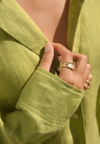 Lniana koszula w jasnym zielonym kolorze z kołnierzykiem i detalem guzików, połączona ze złotym pierścionkiem i subtelną strukturą skóry.