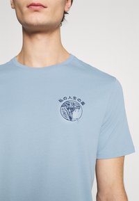 Světle modré bavlněné tričko s krátkým rukávem s tmavě modrým motivem hada v kruhovém designu na levé straně hrudi.