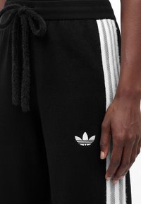 Κοντινό πλάνο μαύρων παντελονιών φόρμας Adidas με λευκές τρεις ρίγες στο πλάι και κεντημένο λογότυπο Adidas trefoil, που δείχνει το χέρι ενός ατόμου να ξεκουράζεται κοντά.