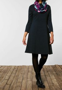 Czarna sukienka w ciemnoniebieskim kolorze z teksturowanym wzorem, z rękawami o długości trzy czwarte i rozkloszowanym dołem, zestawiona z kolorowym, drukowanym szalikiem i czarnymi botkami.