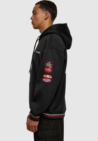Schwarzer Kapuzenpullover mit bestickten Patches am rechten Ärmel, featuring roten und weißen Designs; gerippte Bündchen und Saum, frontale Taschen.