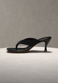 Čierna sandála na vysokom podpätku so štvorcovou špičkou a širokým remienkom, zobrazená na svetlom okrúhlom povrchu na jednoliatom pozadí.