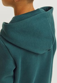 Felpa teal con una texture morbida, dotata di un grande cappuccio piegato. Il tessuto appare leggermente peloso, mettendo in risalto il suo comfort e calore.
