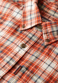 Chemise en flanelle à carreaux rouges et blancs avec des accents bleus et jaunes, dotée de boutons marron et d'une étiquette de logo. Tissus texturé avec une finition douce.