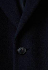 Marineblå uldblandingsfrakke med spidse krave, en enkelt sort knaplukning og en blød, struktureret overflade.