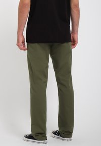 Olijfgroene broek met een rechte pijpen, gemaakt van een glad materiaal. Gecombineerd met zwarte hoge sneakers. Geen opvallende patronen of accenten.