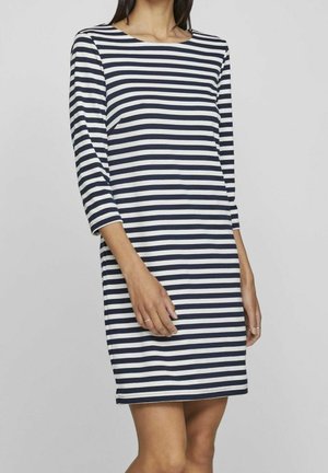 Femme portant une robe aux rayures horizontales bleu marine et blanches, longueur au genou, avec des manches trois-quarts, sur un fond gris uni.