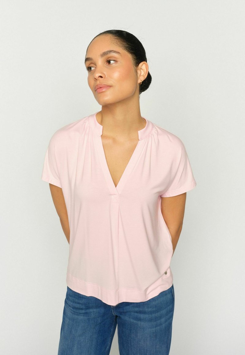 Femme aux cheveux foncés attachés en chignon, portant un chemisier rose clair à manches courtes et col en V, ainsi qu'un jean bleu, debout devant un fond clair uni.