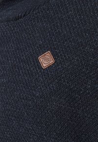 Tissu tricoté bleu foncé texturé avec une petite pièce en cuir marron en forme de losange portant l’étiquette « Springfield », cousue près du centre.
