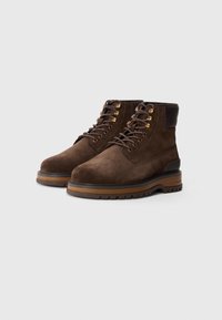 Botas de ante marrón con cordones, ojales de metal y suelas de goma marrón. Presentan un upper texturizado con un cuello de cuero oscuro en contraste.