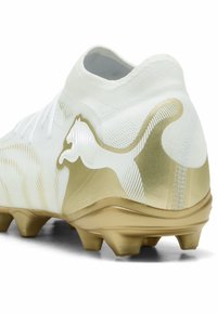 Λευκό και χρυσό ποδοσφαιρικό παπούτσι με διαμορφωμένες τάπες και στυλιζαρισμένο λογότυπο αλματώδους πυμά στη πλευρά κοντά στη φτέρνα.