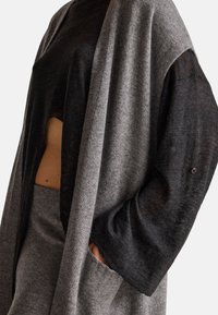 Šedý a černý pletený top s širokými rukávy, zkrácený střih odhalující břicho a detail knoflíku na rukávu. Měkká textura a vrstvený vzhled.