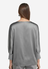 Blusa in satin grigio con una vestibilità rilassata, maniche a 3/4 con palloncino e scollatura ampia. La texture liscia e la leggera lucentezza esaltano il design.