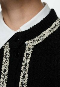 Czarny kardigan o teksturowanej powierzchni z kremowym wzorem dekoracyjnym z przodu. Posiada wysoki kołnierz oraz czarne guziki do zapięcia.