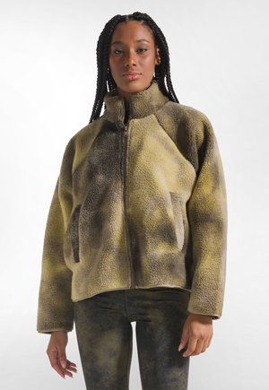 Vrouw met lange vlechten die een bruine en groene fleecejas met ritssluiting en bijpassende legging draagt, staand tegen een effen achtergrond.