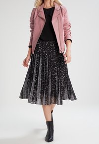 Giacca motociclista in suede rosa chiaro sopra una blusa nera e una gonna plissettata nera con macchie bianche. Indossata con stivaletti neri.