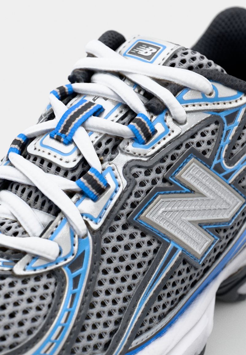 Grijze sportieve schoen met een bovenwerk van mesh, voorzien van blauwe en zilveren accenten, witte veters en een groot zilveren "N"-logo aan de zijkant.