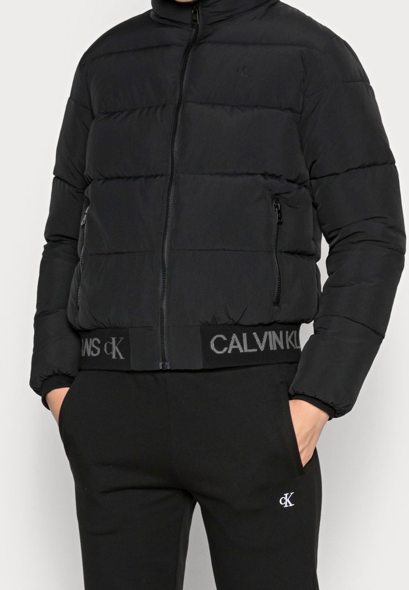 Giubbotto puffa nero con cuciture orizzontali, colletto alto, zip anteriore e tasche laterali. Presenta una cintura elastica con logo "CALVIN KLEIN."