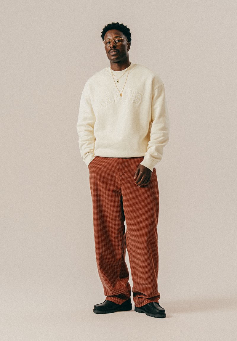 Mężczyzna w okularach, ubrany w kremowy sweter, czerwone spodnie w kratę, czarne buty i dwa złote naszyjniki, stoi na beżowym tle.