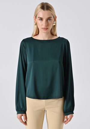 Blusa lunga in satin teal con scollatura a barca, vestibilità rilassata e polsini arr gathered; abbinata a pantaloni aderenti beige chiaro.