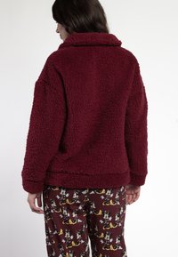 Chaqueta de piel sintética burdeos con una superficie texturizada y esponjosa y un cuello doblado. Combinada con pantalones de pijama estampados que presentan personajes de dibujos animados.