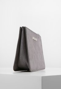 Šedá semišová kabelka přes rameno s jednoduchým obdélníkovým tvarem, která má na přední straně stříbrný kovový detail a zapínání na zip.