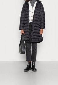 Zwarte donsjas met capuchon, voorzien van een doorgestikt ontwerp. Draagt over een grijze trui met knopen, gecombineerd met een zwarte jeans en enkellaarsjes.