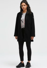 Black fleece coat with a hood, featuring a full zipper, long sleeves, and side pockets, layered over a light graphic t-shirt and black pants.
