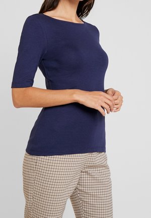 Marineblå tætsiddende T-shirt med bådhals og ærmer til albuen, lavet af et glat stof, parret med ternede brune bukser.