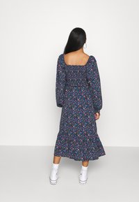 Kukallinen, pitkähihaine mekko, jossa on sininen pohja ja vaaleanpunaisia ja vihreitä kukkia, neliskulmainen kaula-aukko, ryppyinen vartalo ja röyhelöhimo.