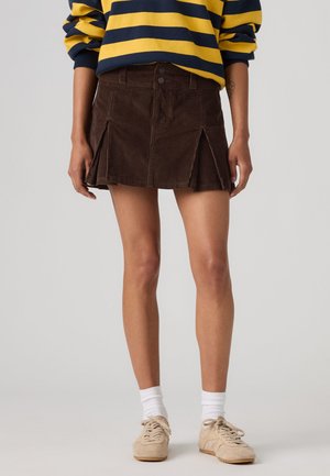 Persona che indossa una minigonna in velluto a coste marrone con pieghe frontali, una maglia a maniche lunghe gialla e blu navy a righe, calzini bianchi e sneaker beige.
