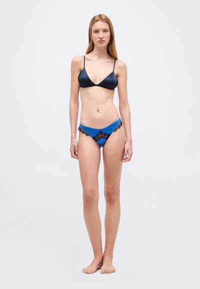 Γυναίκα που στέκεται ξυπόλυτη, φοράει σκούρο μπλε τριγωνικό σουτιέν και μπλε σλιπ με μαύρη δαντέλα, μπροστά σε απλό φωτεινό φόντο.