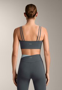 Tummanharmaa urheiluliivit ohuilla olkaimilla ja logolla, yhdistettynä korkeavyötäröisiin leggingseihin, joissa on vaaleanharmaa korostus vyötäröllä. Sileä kangaspinta.