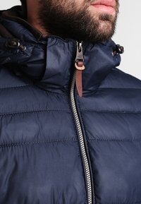 Chaqueta acolchada azul marino con collar alto, cierre de cremallera y tirador con acentos de cuero; presenta costuras horizontales y un acabado resistente al agua.