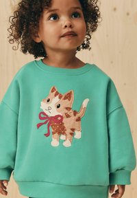 Felpa turchese con un'applicazione di un gatto in peluche con un fiocco rosa. Il design include dettagli come macchie e una texture ricciosa.