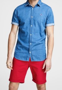 Hombre con una camisa de mezclilla de mangas cortas y pantalones cortos rojos, de pie con las manos relajadas a los lados, mostrando un atuendo casual de verano.