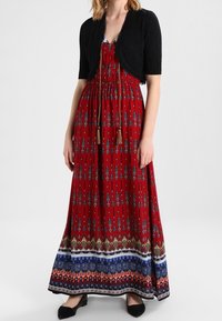 Czerwona sukienka maxi z niebiesko-białymi wzorami, zestawiona z czarnym krótkim kardiganem. Posiada talię ze sznurkiem i ozdobne frędzle.