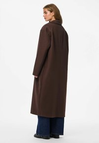Lungo cappotto di lana marrone con colletto e taglio dritto, caratterizzato da maniche lunghe e una trama semplice, abbinato a jeans a gamba ampia.
